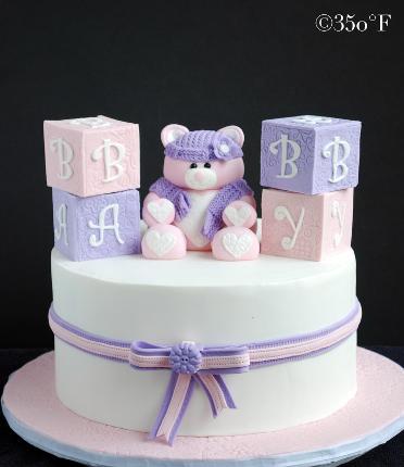 A baby shower cake with edible baby blocks and a teddy bear welcoming a baby girl.